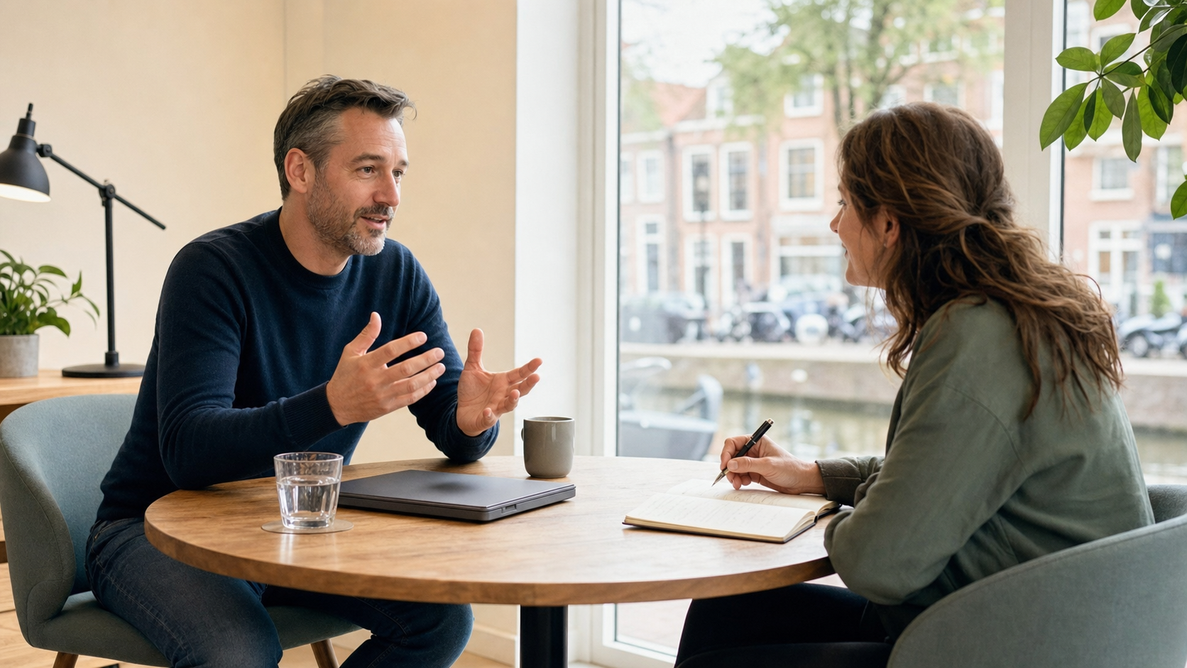 Rustig eerste gesprek aan een ronde tafel met notitieboek, koffie en uitzicht op een Nederlandse straat